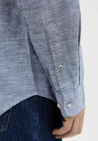 Cămașă din in bleu deschis, cu manșete cu două nasturi, tiv drept și textură subtilă, purtată peste blugi din denim închis.