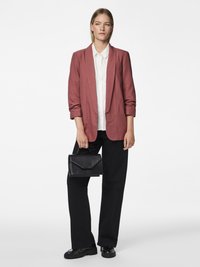 Femme portant un blazer rose, une chemise blanche boutonnée, un pantalon large noir, des mocassins noirs, tenant un sac à main noir, debout devant un fond blanc.