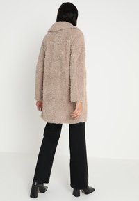 Beżowy płaszcz z sztucznego futra z szerokim kołnierzem, długimi rękawami i luźnym krojem, zestawiony z czarnymi spodniami i botkami za kostkę.