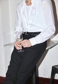 Balta pogu krekls pār balto bezdžemperi, melni bikses, tur rokās vīna glāzi. Marmora galds un melnais krēsls fona.