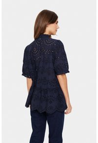 Marineblaue Bluse mit Lochstickerei, rüschigen kurzen Ärmeln und einem gestuften Saum. Der Stoff hat eine weiche Textur und filigrane Muster.