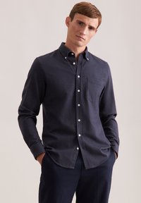Giovane uomo che indossa una camicia button-up blu scuro con taschino, in piedi con le mani in tasca su uno sfondo chiaro e semplice.