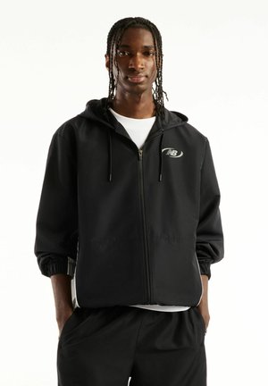 Ung mand med flettet hår iført sort hoodie med hvid New Balance-logo og sorte bukser, hænderne i lommerne, vendt fremad.
