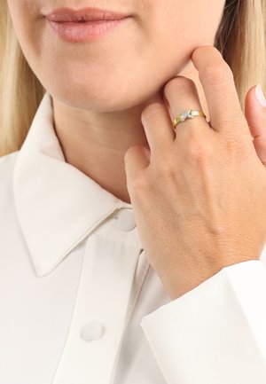 Anello d'oro con accento in argento caratterizzato da un design floreale, indossato su una mano con pelle chiara, su uno sfondo di camicia bianca con colletto.