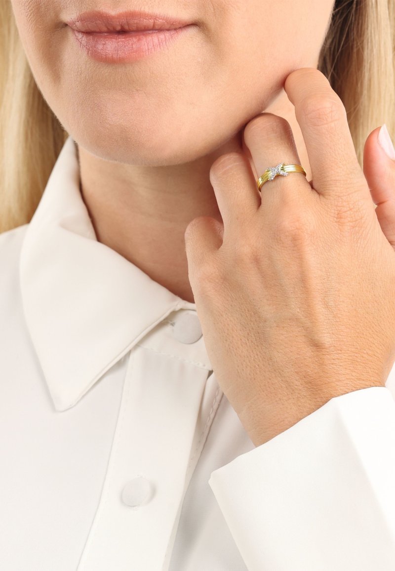 Złoty pierścionek z srebrnym akcentem, zdobiony wzorem kwiatowym, noszony na dłoni o jasnej karnacji, na tle białej koszuli z kołnierzykiem.