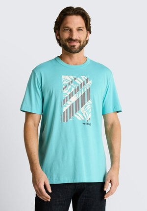 Mężczyzna w jasnoniebieskim T-shircie z geometrycznym paskowanym i liściastym wzorem oraz czarnych spodniach, stojący na jednolitym tle.