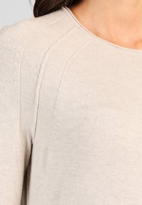 Maglione beige chiaro con una vista ravvicinata della sua texture lavorata a maglia e dei dettagli delle cuciture, che evidenziano uno stile casual e accogliente.