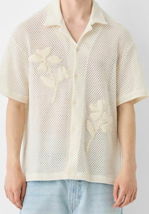 Beige mesh kortærmet skjorte med knapper og krave med blomsterbroderi, båret med lyseblå jeans.