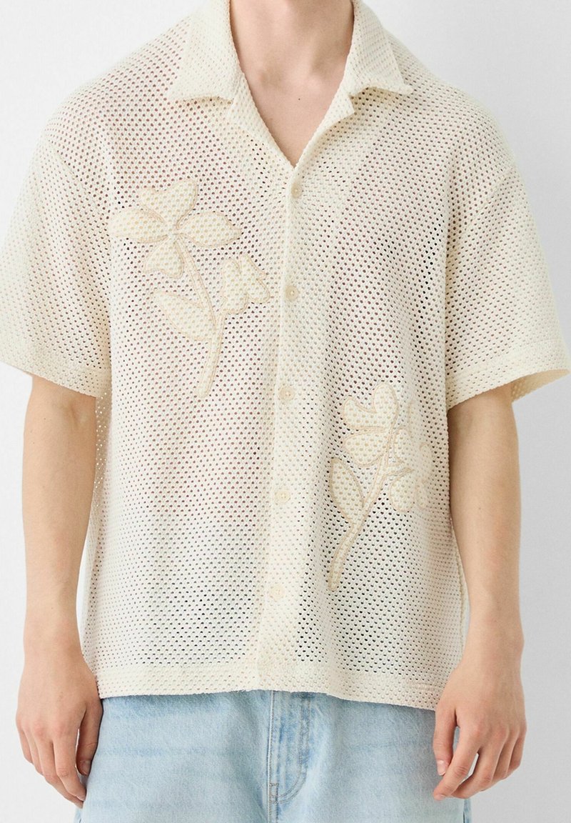 Chemise beige en maille à manches courtes, boutonnée, avec col et broderie florale, portée avec un jean en denim bleu clair.