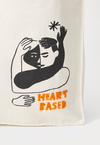Canvas tote bag featuring black line art of intertwined hands and faces. Text reads "HEART BASED" in orange embroidery.