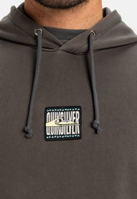 Grå hettegenser med en teksturert overflate, med et fremtredende Quiksilver-logo-patch og justerbare snorer på hetten.