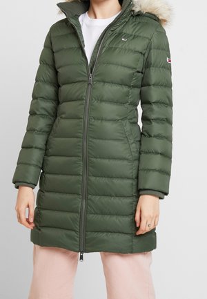 Femme portant un long manteau d'hiver matelassé vert olive avec capuche bordée de fourrure et fermeture éclair à l'avant, assorti à un pantalon rose clair.