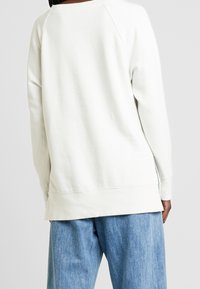 Sudadera blanca con un corte holgado, mangas raglán y un dobladillo trasero ligeramente más largo. Material suave y texturizado. Combinada con pantalones de mezclilla azul claro.