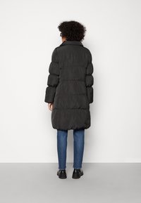 Black puffer coat with a long silhouette, quilted texture, wide sleeves, and a collar. Worn with blue jeans and black shoes.