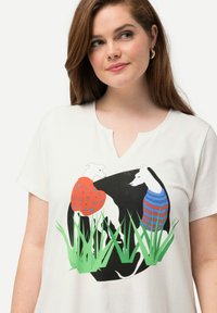 Camiseta blanca de algodón con un diseño gráfico de dos perros vestidos con prendas coloridas sobre un fondo redondo negro con acentos de hierba verde.