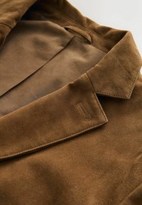 Brun ruskindsjakke krave med en glat tekstur, der har synlige sømme og et diskret præget logo detalje på stoffet.