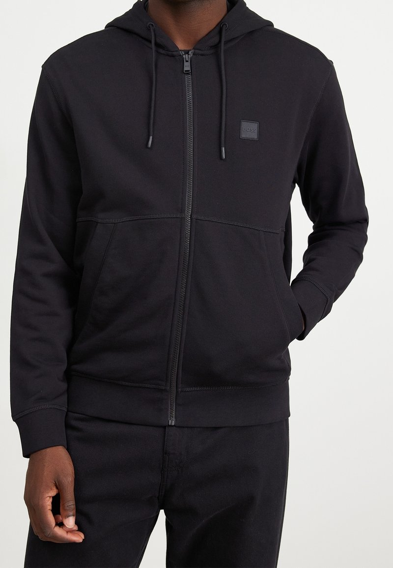 Homme portant un sweat à capuche noir zippé avec poches avant et un petit logo carré "BOSS" sur le côté gauche de la poitrine sur un fond uni.