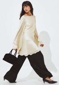 Dolga majica kremaste barve z ohlapnim krojem, v kombinaciji z širokimi črnimi hlačami in črnimi salonarji z ostrimi konicami, drži majhno črno torbico.