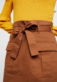 Gonna di cotone marrone con cintura annodabile, due tasche frontali e una forma strutturata. Il materiale ha una texture liscia.