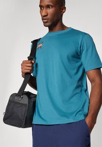 Teal katoenen T-shirt met korte mouwen en een ronde halslijn, voorzien van een oranje grafisch detail. Zwarte tas met een gestructureerde zijkant en een verstelbare band.