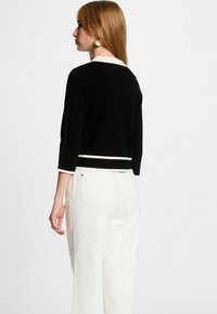 Zwarte cropped trui met witte afwerking, driekwart mouwen, gemaakt van zachte stof. Model draagt het met een hoge taille, witte wijde broek.