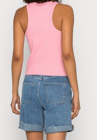 Kobieta w różowej bluzce bez rękawów i skróconych niebieskich dżinsowych szortach, przedstawiona od tyłu na jednolitym jasnym tle.