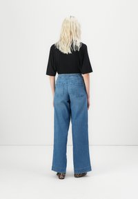 Weite blaue Jeans aus Denim mit hoher Taille, Gesäßtaschen und einer glatten Oberfläche, kombiniert mit einem kurzen schwarzen Oberteil und gemustertem Schuhwerk.