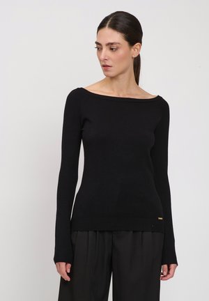 Frau mit dunklem, zurückgebundenem Haar, die ein eng anliegendes schwarzes Langarmshirt und lockere schwarze Hosen trägt, schaut leicht nach links vor weißem Hintergrund.