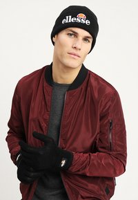 Bordový bomber kabát s vroubkovaným límcem, černá čepice s bílým logem a černé rukavice s logem. Model má neutrální výraz.