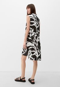 Ärmelloses schwarz-weißes Kleid mit einem Blättermuster, Leinenstruktur, knielangem Design und seitlichen Schlitzen. Trägt schwarze Sandalen.
