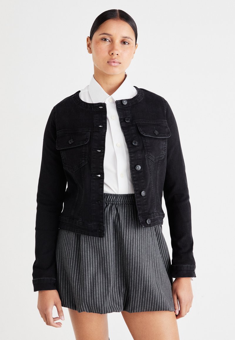 Μαύρο τζάκετ από τζιν με μακριά μανίκια, μπροστινά κουμπιά και δύο τσέπες στο στήθος. Φορεμένο πάνω από ένα λευκό πουκάμισο με γιακά, συνδυασμένο με ριγέ σορτς.