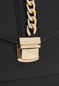 Czarna skórzana torebka z złotym łańcuszkowym paskiem i prostokątnym złotym zapięciem o fakturowanej powierzchni. Gładka powierzchnia z przeszyciami jako detalami.