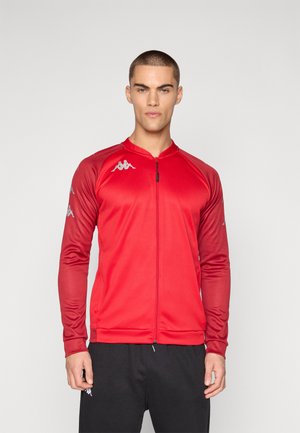 Modello maschile che indossa una giacca sportiva rossa con zip e loghi sulle spalle e le maniche, abbinata a pantaloni neri, in piedi davanti a uno sfondo semplice.