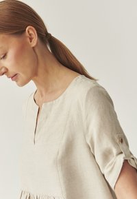 Robe en lin beige doux avec un col rond et une taille froncée. Présente des manches à revers avec des boutons et une texture subtile.