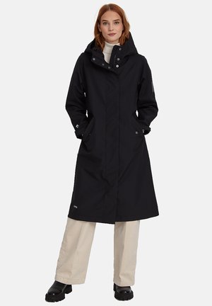 Kobieta o rudych włosach, ubrana w czarny długi płaszcz z kapturem, beżowe szerokie spodnie i czarne masywne buty, stoi z rękami w kieszeniach.