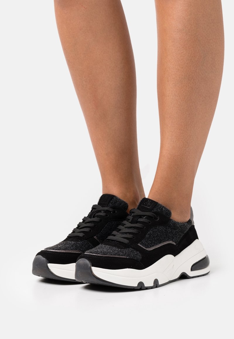 Baskets athlétiques noires et grises avec une tige en tissu texturé, des accents en daim et une semelle blanche épaisse. Dotées de lacets ronds et d'un col rembourré.