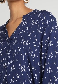 Navy blouse with a lightweight fabric, featuring a white bird pattern. V-neckline and button front detailing. Textured, relaxed fit.