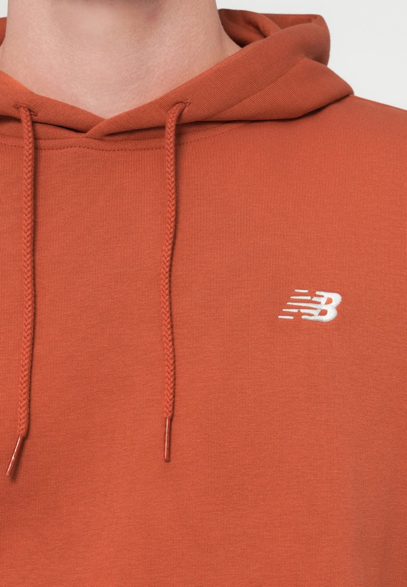 Hoodie in gebranntem Orange mit einer vorderen Kängurutasche. Verfügt über ein weißes, gesticktes Logo auf der Brust und verstellbare Kordeln. Glatte Textur.