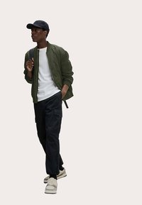 Olivengrøn bomberjakke, hvid T-shirt, sorte cargobukser og lyse sneakers. Modellen bærer en sort kasket og står foran en neutral baggrund.