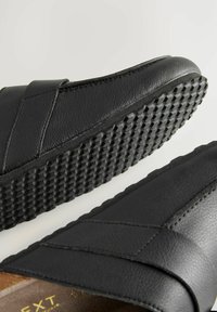Crne kožne slip-on cipele s teksturiranom potplatom i dizajnom ukriženih remena. Gornji deo ima glatku obradu i vidljive detalje šivenja.