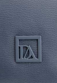 Cuero texturizado de color azul oscuro con un logo cuadrado elevado que presenta las letras "DA" en una tipografía estilizada, centrado en la superficie.