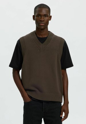 Homme portant un pull sans manches en tricot marron par-dessus une chemise noire à manches courtes, une main dans la poche du jean, debout devant un fond uni.