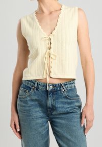 Krémový pletený crop top s hlubokým V-výstřihem, klokaními okraji a předním zavazováním. V kombinaci s vysokými modrými džínami.