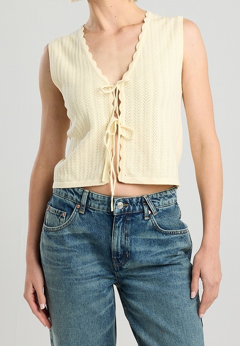 Krémový pletený crop top s hlubokým V-výstřihem, klokaními okraji a předním zavazováním. V kombinaci s vysokými modrými džínami.