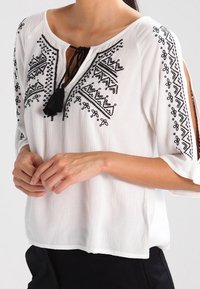 Hvid bluse med sorte broderede mønstre, korte ærmer og en bindesløjfe ved halsen med kvastdetalje. Letvægtsstof med let tekstur.