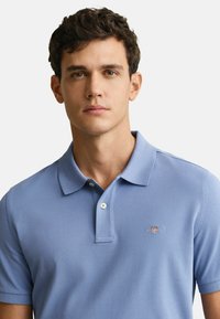 Pikeepaita vaaleansinisestä kankaasta, jossa on röyhelökaulus ja kolmonen nappi. Vasemmassa rinnassa on pieni logo. Lyhyet hihat.