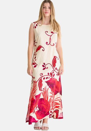 Femme portant une robe maxi crème sans manches avec de grands motifs floraux rouges et des feuilles, associée à des talons blancs ouverts, debout devant un fond uni.
