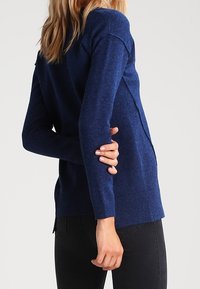 Kobieta ubrana w dopasowany granatowy sweter i czarne dżinsy, z jedną ręką zgiętą za plecami trzymającą przeciwny łokieć.