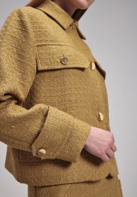 Giacca gialla testurizzata con accenti di bottoni dorati, due taschini sul petto e un design del colletto classico. Dettaglio ravvicinato della manica mostrato.