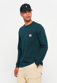 Lange mouwen teal shirt met een borstzak en een vierkante logo patch, gecombineerd met tan cargobroeken en een zwarte pet met een logo.